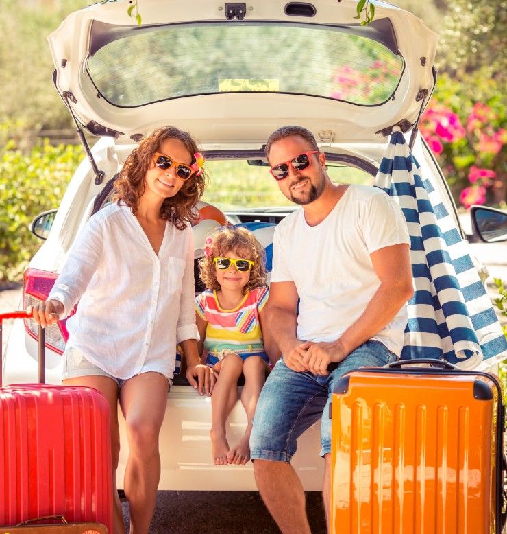 Mom, dad and their daughter pose with red and orange suitcases and beach gear on a tropical vacation. 